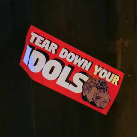 red rectangular sticker decal on a lamp post with the words "tear down your idols" in white all-caps with black shadow and an illustration of the statue of John A MacDonald's statue in the foreground