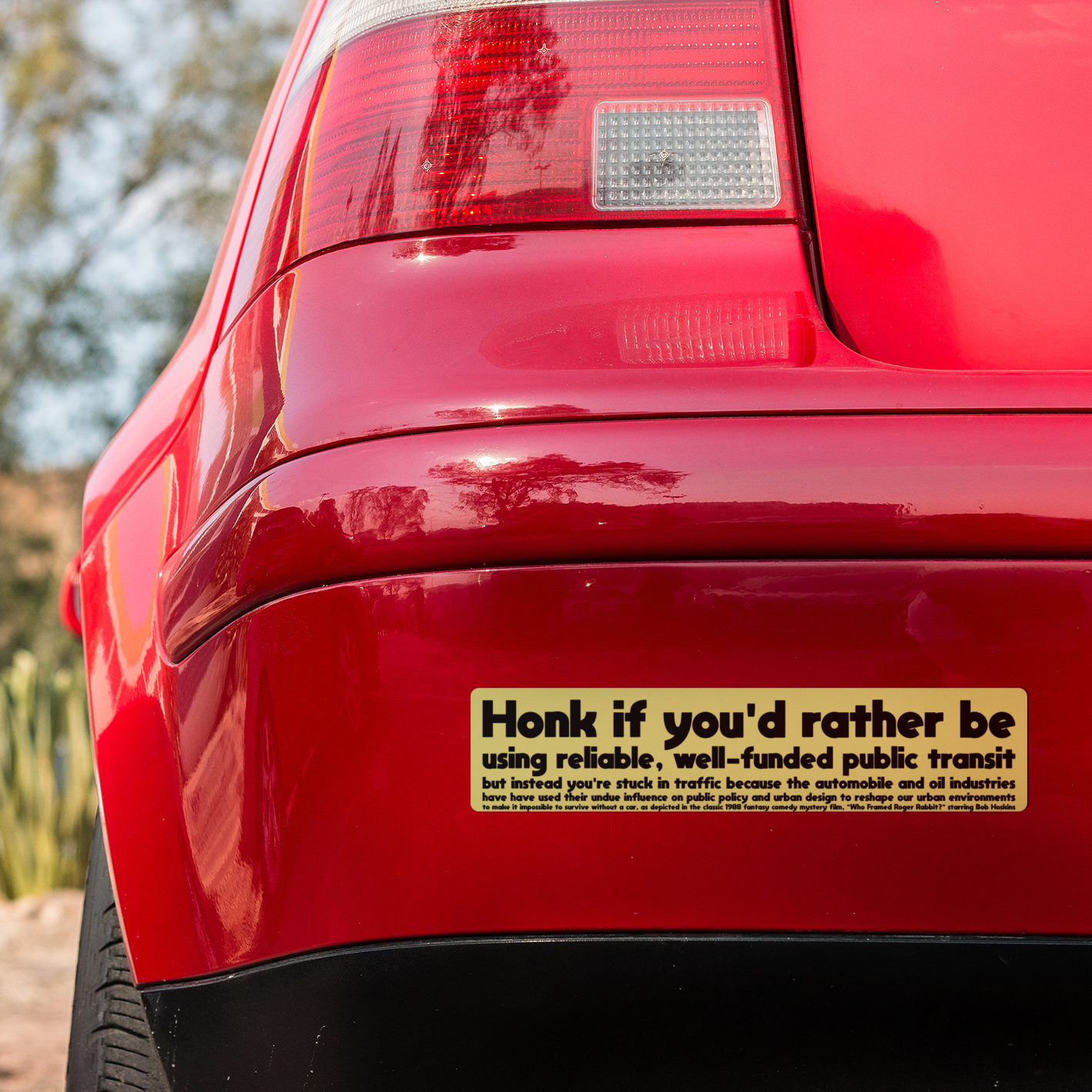 Red car bumper with yellow bumper sticker reading " "Honk if you'd rather be using reliable, well-funded public transit but instead you're stuck in traffic because the automobile and oil industries have used their undue influence on public policy and urban design to reshape our urban environments to make it impossible to survive without a car, as depicted in the classic 1988 fantasy comedy mystery film. "Who Framed Roger Rabbit, starring Bob Hoskins"