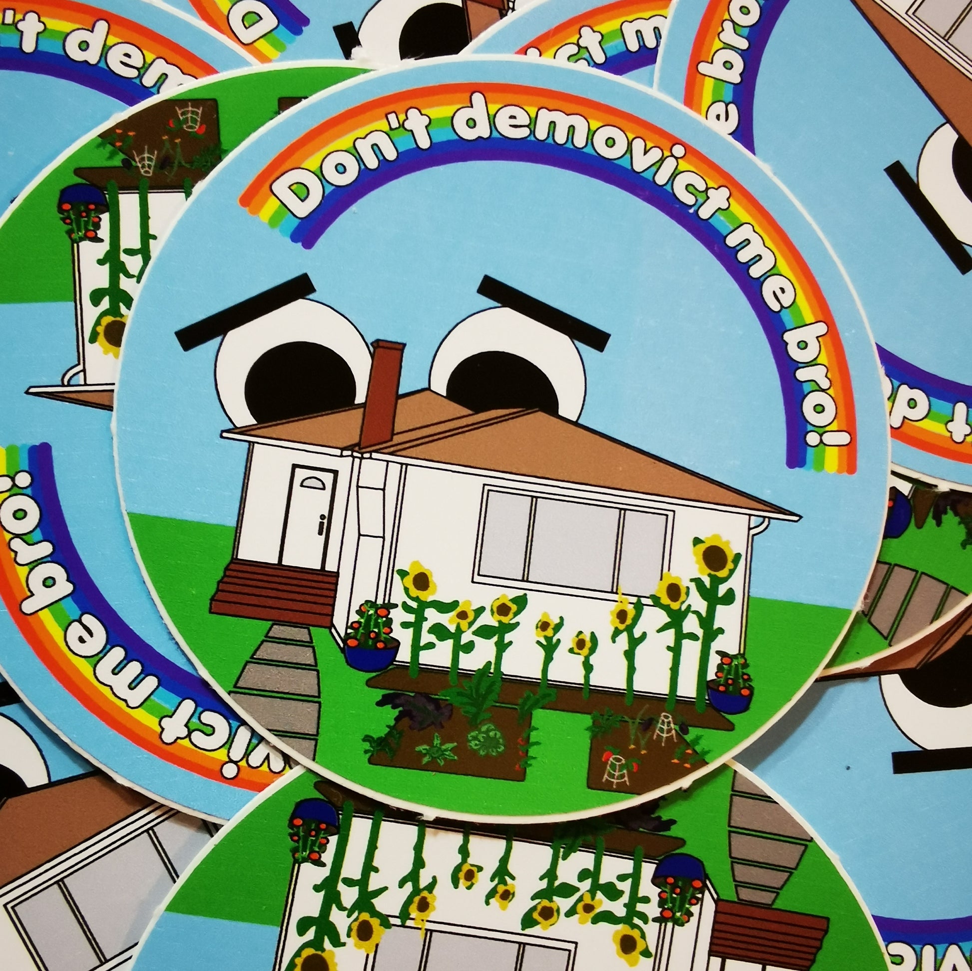 circle sticker with white detached home with sunflowers and a vegetable garden. The house has big cartoon eyes on the roof and worried eyebrows. there is a rainbow in the sky with the words "don't demovict me, bro!"