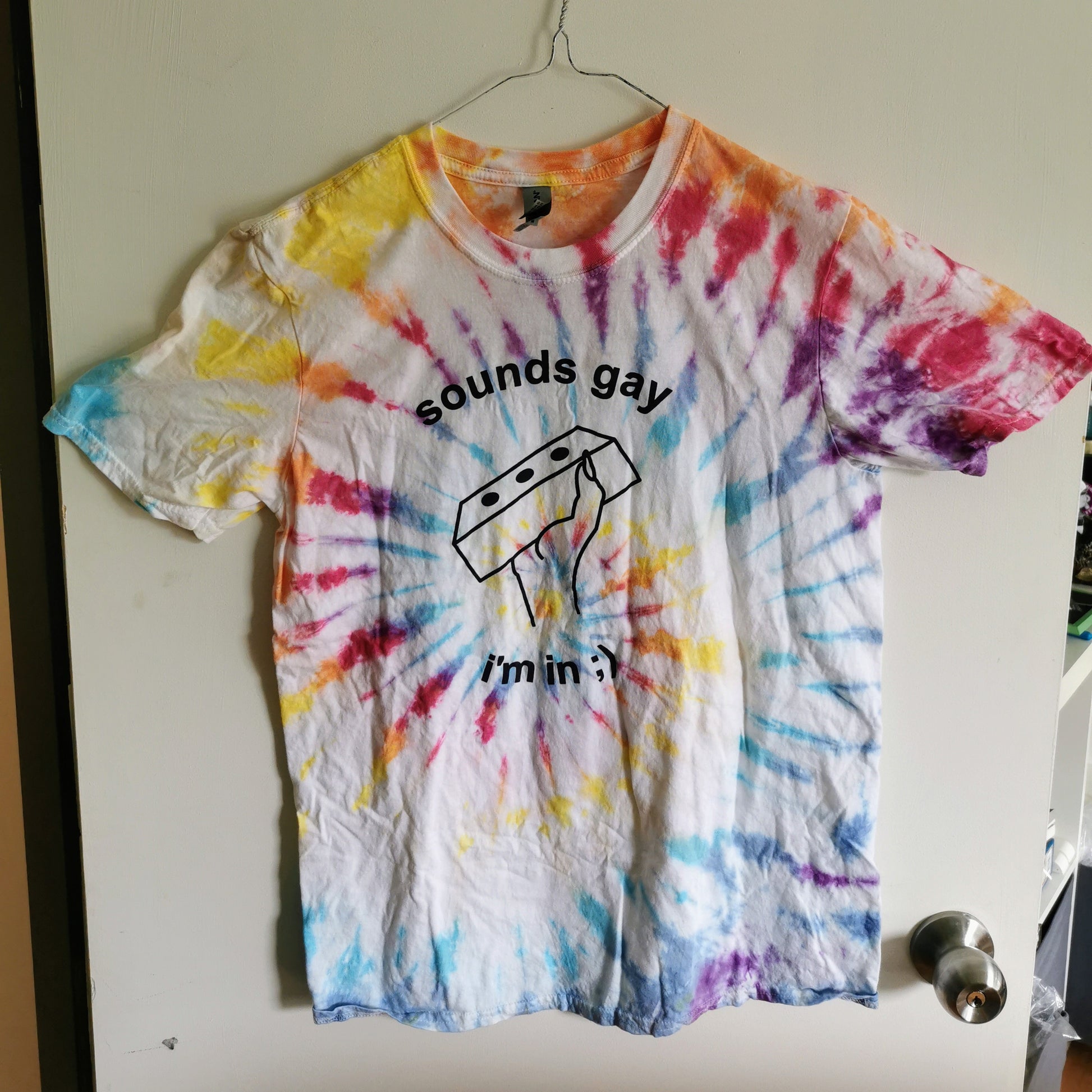 rainbow swirl tie-dye t-shirt with text reading "sounds gay i'm in" and a line drawing of a hand holding a brick