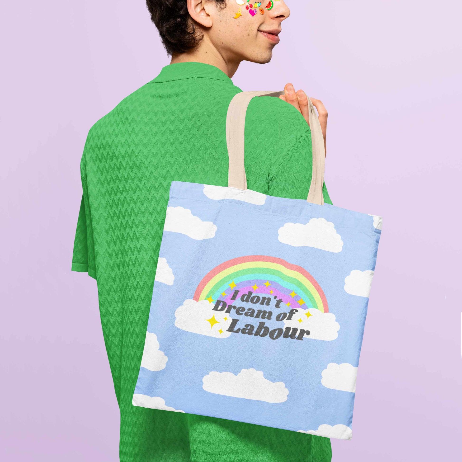 Person wearing bright green collared button down herringbone shirt with fruit stickers on their face with a blue tote bag with clouds, a rainbow and the words I don't dream of labour surrounded by yellow sparkle icons