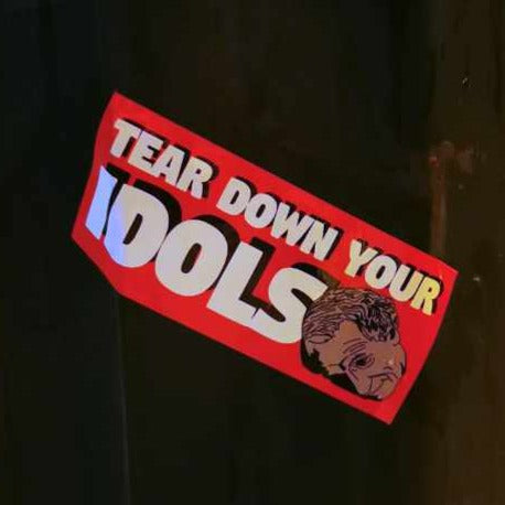 red rectangular sticker decal on a lamp post with the words "tear down your idols" in white all-caps with black shadow  and an illustration of the statue of John A MacDonald's statue in the foreground