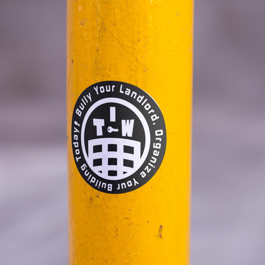 black and white circle sticker with the words "bully your landlord. Organize your building today!" around the edge and an apartment building in a circle with the letters "I T W" above with a key