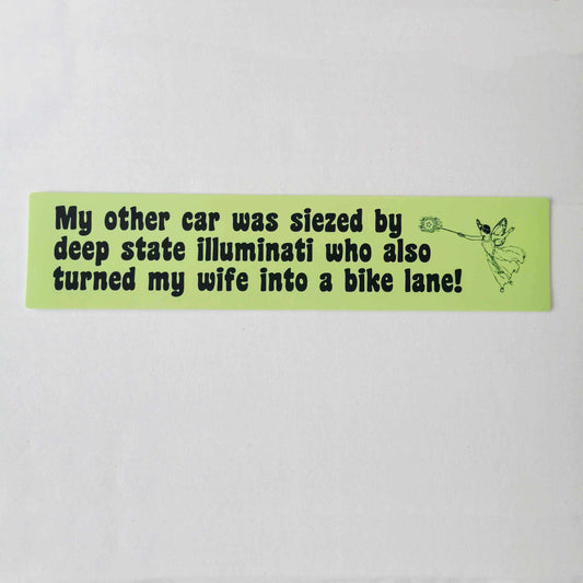 Light green bumper sticker with chunky curvy text reading "My other car was seized by deep state illuminati who also turned my wife into a bike lane!" and features an old-timey fairy with a magic wand who works for the deep state and is definitely the one who will turn your wife into a bike lane.