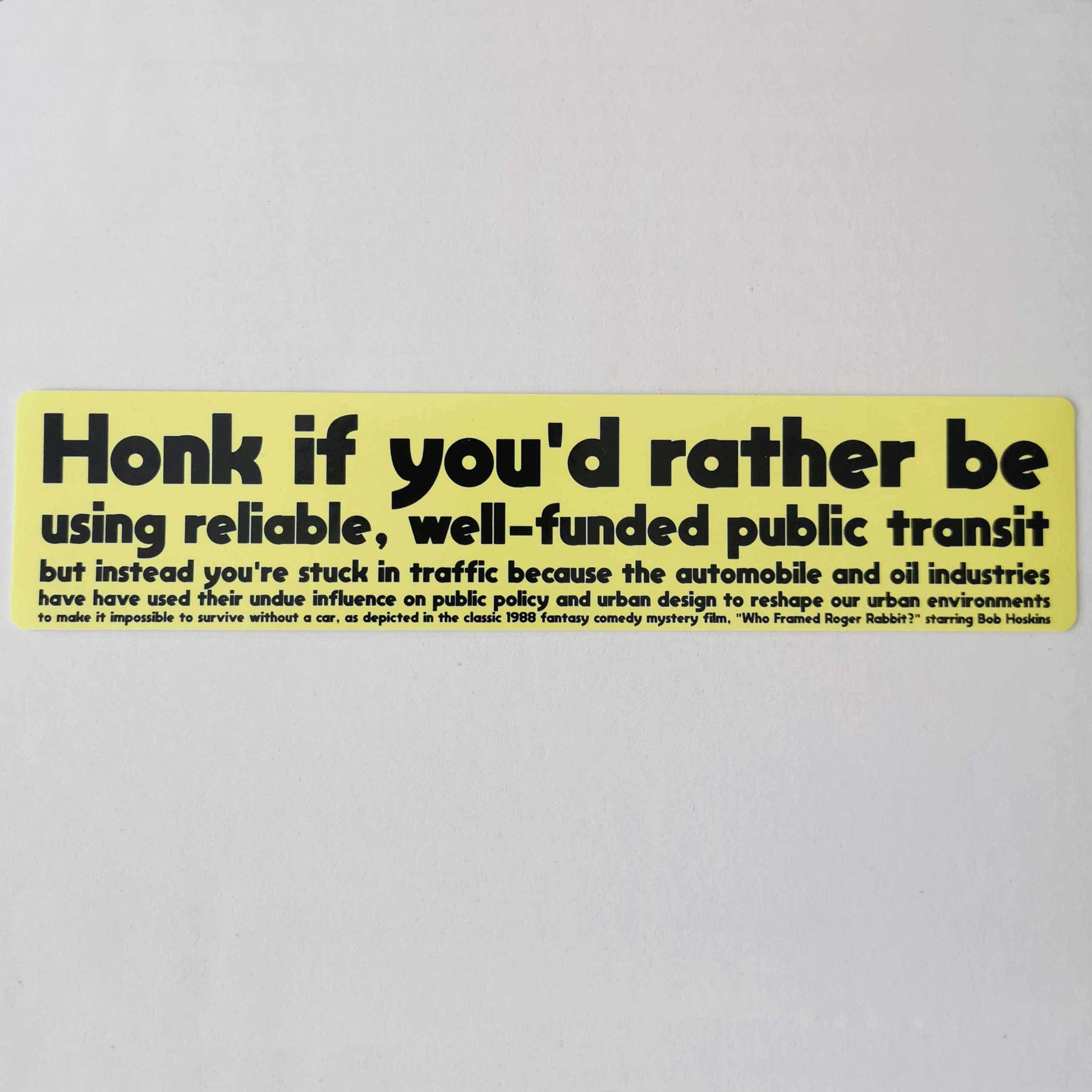 Light yellow bumper sticker reading ""Honk if you'd rather be using reliable, well-funded public transit but instead you're stuck in traffic because the automobile and oil industries have used their undue influence on public policy and urban design to reshape our urban environments to make it impossible to survive without a car, as depicted in the classic 1988 fantasy comedy mystery film. "Who Framed Roger Rabbit, starring Bob Hoskins" in bold black sans serif font that gets smaller with each line