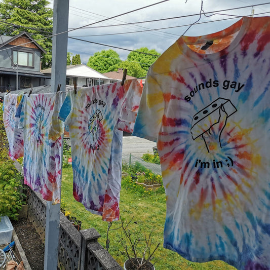 rainbow swirl tie-dye t-shirts with text reading "sounds gay i'm in" and a line drawing of a hand holding a brick. shirts are hung on a clothesline outside