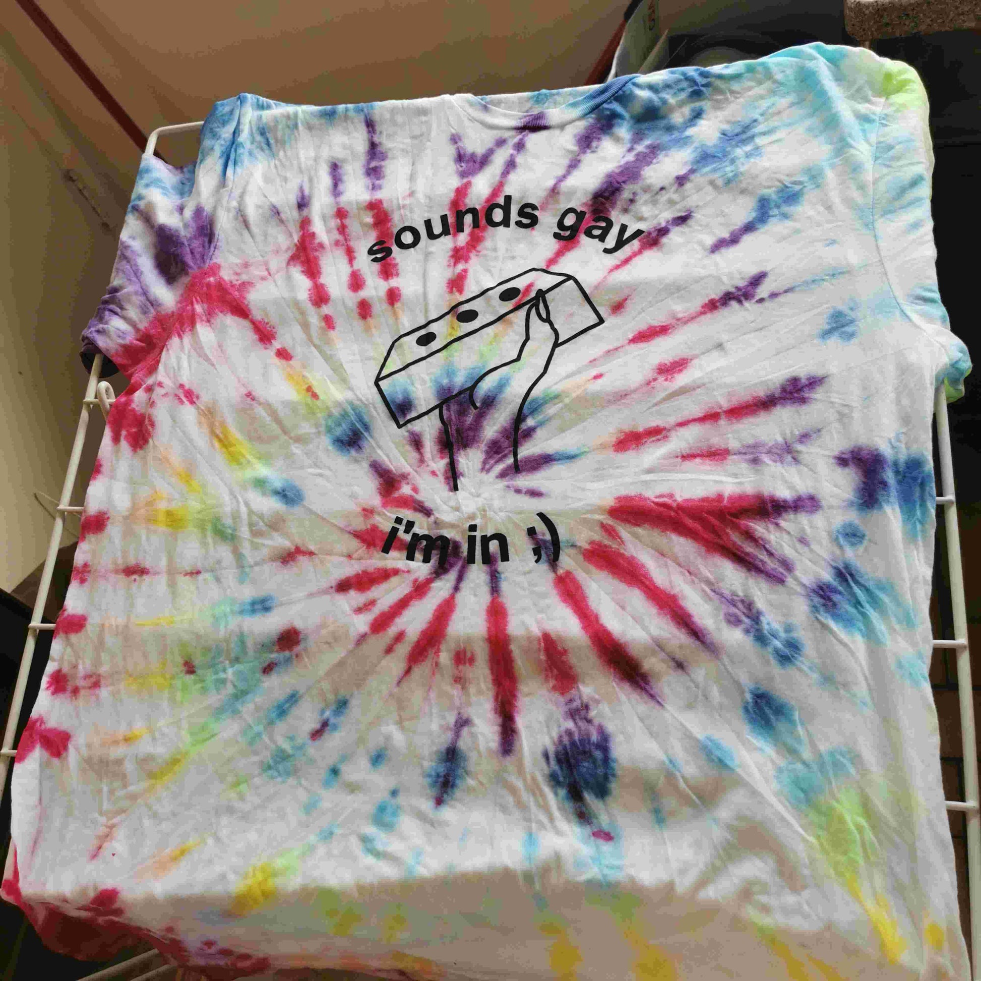 rainbow swirl tie-dye t-shirts with text reading "sounds gay i'm in" and a line drawing of a hand holding a brick. Shirt is drying on a drying rack
